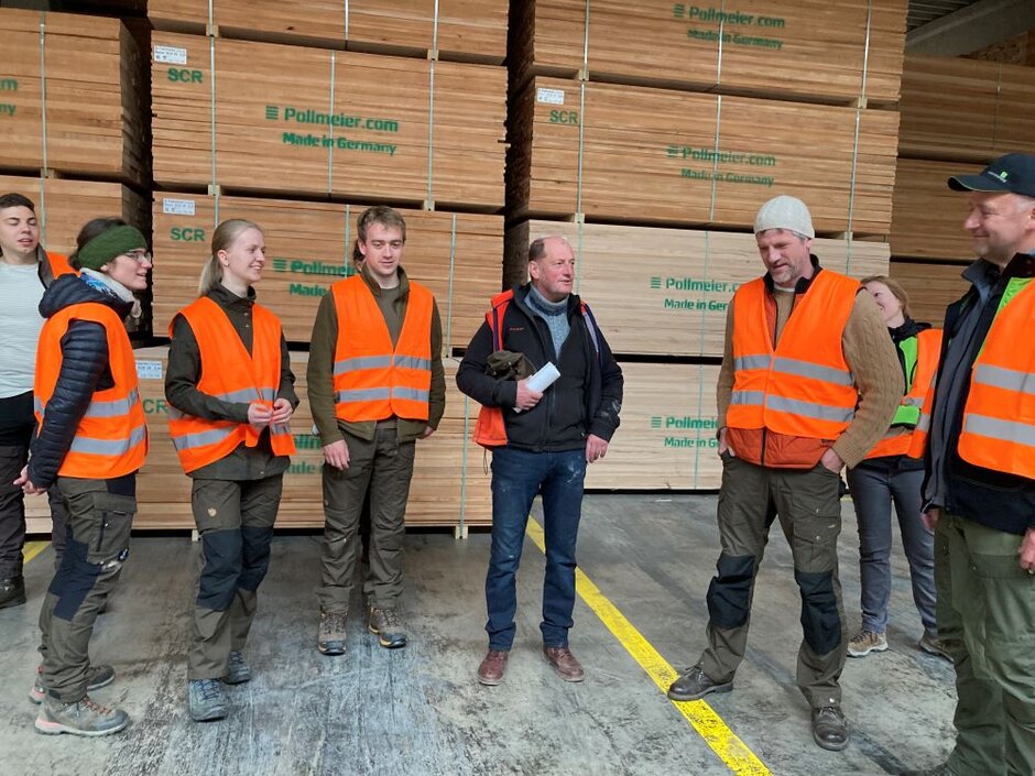 Die Studenten nehmen an einer Führung durch das Sägewerk teil. Sie stehen von gestapelten Holzbrettern.