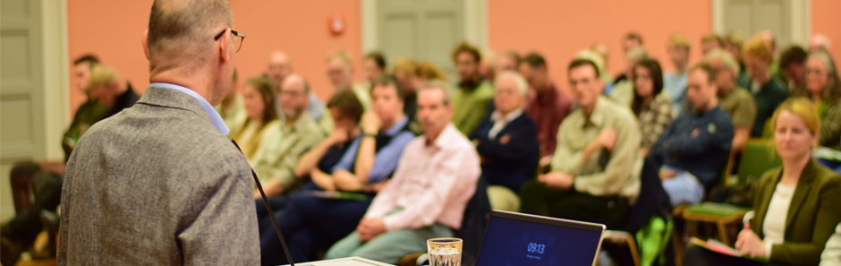 Ein Redner in einem Jackett spricht vor einem forstfachlichen Publikum. Vor ihm steht ein aufgeklapptes Laptop