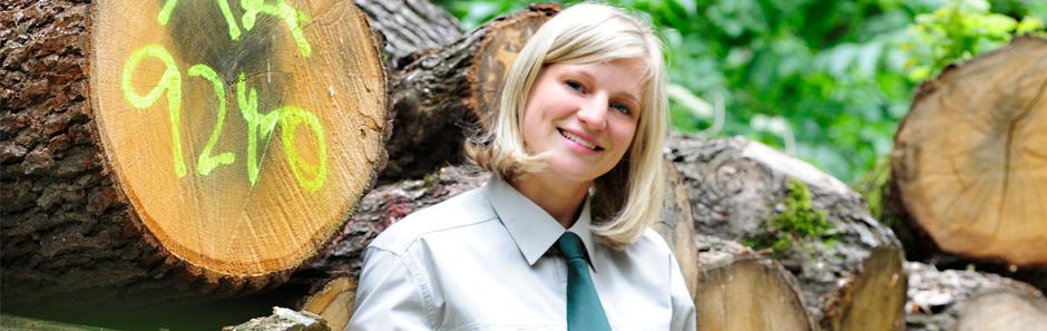 Junge Frau in forstlicher Dienstkleidung steht vor einem Holzpolter im Wald