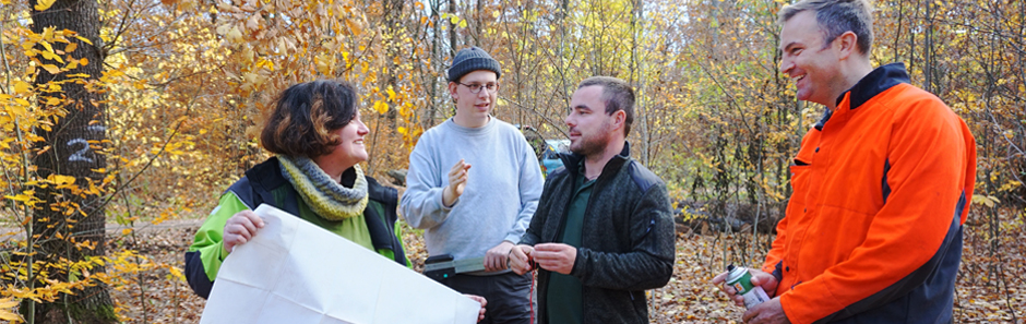 Försterin mit Übersichtskarte in der Hand unterhält sich mit drei interessierten Praktikanten im Herbstwald.