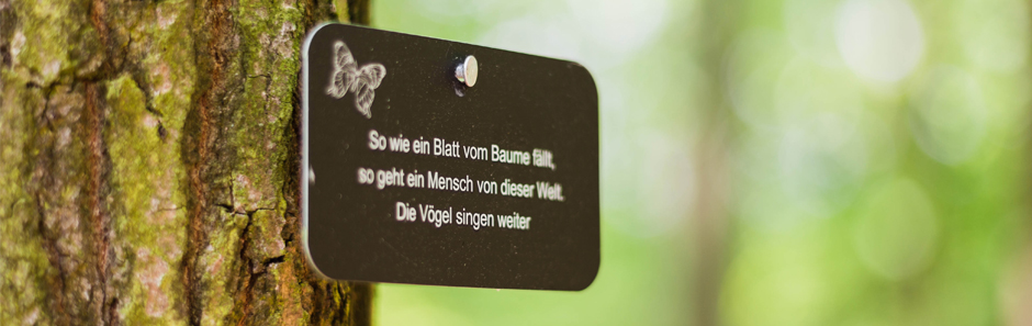 Marke hängt an einem Baumstamm. Auf der Marke steht dem Spruch: "So wie ein Blatt vom Baume fällt, so geht ein Mensch von dieser Welt. Die Vögel singen weiter."